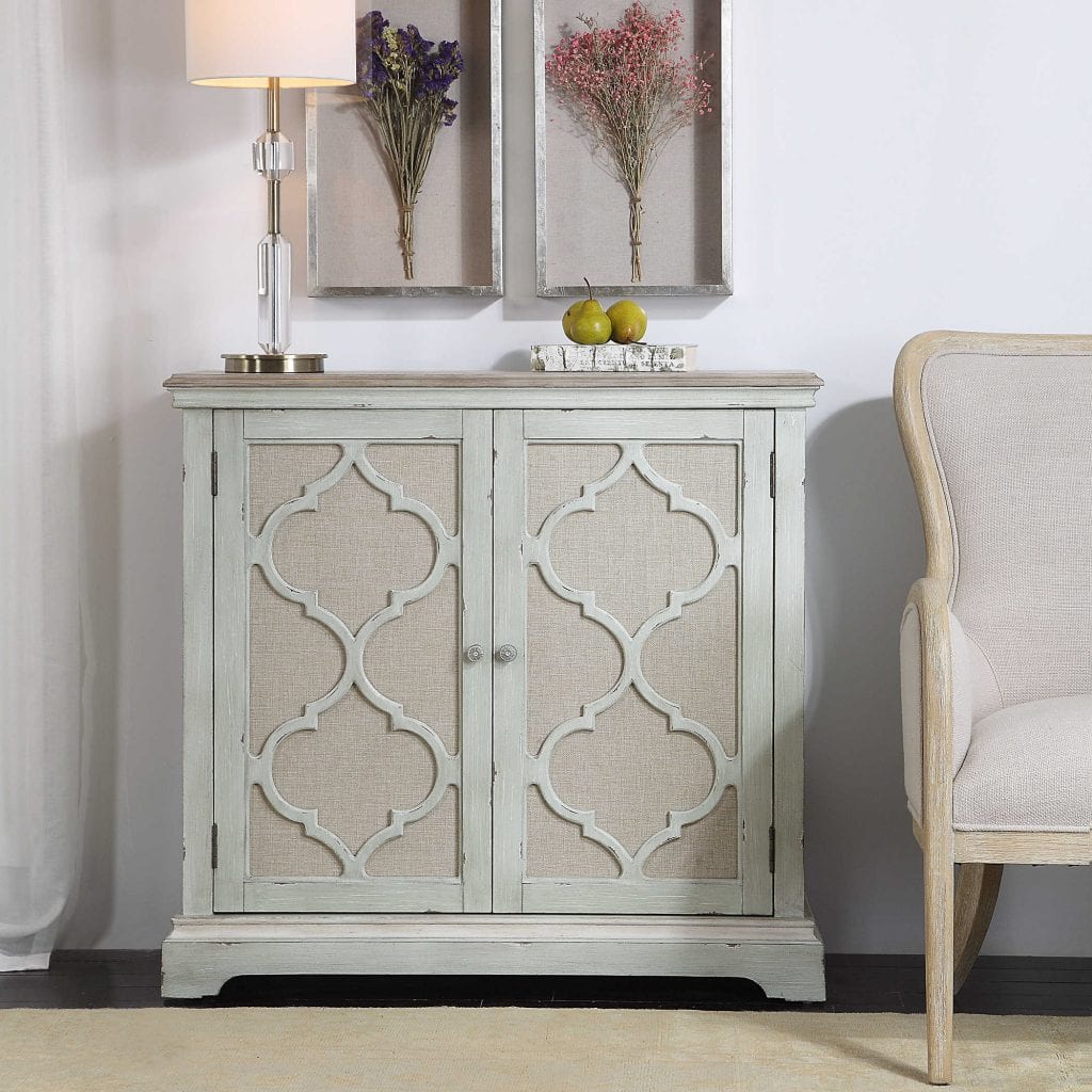 This casual accent cabinet is finished in a weathered sea grey with ivory accents. Open-carved doors are backed in light tan linen, enclosing one adjustable shelf and two storage drawers.